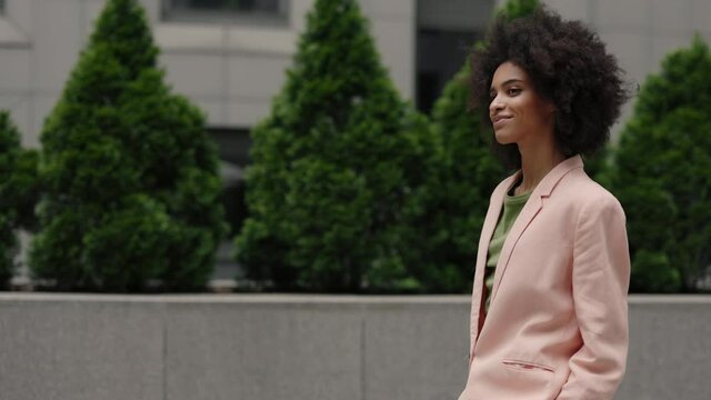 Motion camera view of the happy multiracial businesswoman wearing formal suit walking through the summer street and waving to somebody while being at great mood.