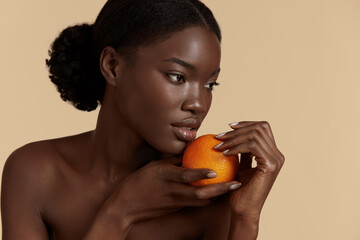 Portrait close up of beautiful african girl hold whole orange. Serious young woman with perfect skin. Concept of skincare. Isolated on beige background. Studio shoot