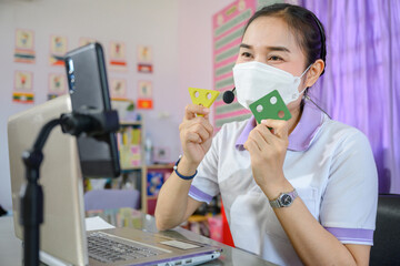 Asian female teacher wearing a mask is teaching students to study online through a computer screen by using an online video conferencing system for education