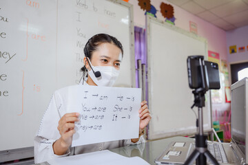 Asian female English teacher wearing a mask teaching students to study online on a computer screen...
