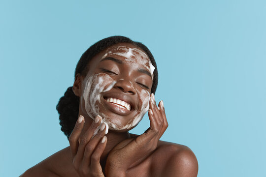 Close up portrait of beautiful black girl wash her face with cleansing face foam. Smiling young woman with closed eyes. Concept of face skin care. Isolated on blue background. Studio shoot