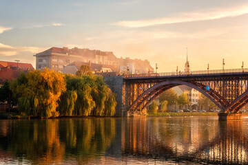 Fototapeta na wymiar Amazing view of Maribor Old city, Main bridge (Stari most) on the banks of Drava river at sunrise, Slovenia. Scenic cityscape with blue sky, morning mist and reflection, outdoor travel background