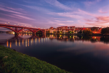 Obraz premium Amazing view of Maribor Old city, Main bridge (Stari most) on the banks of Drava river before sunrise, Slovenia. Scenic cityscape with dramatic cloudy sky and reflection, outdoor travel background