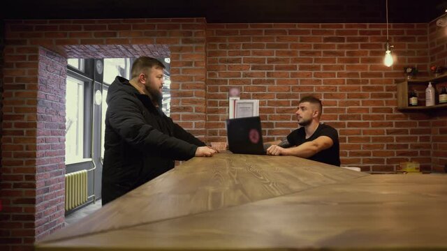 Inside barbershop. Fat chubby client is talking to salon's owner sitting at reception desk. Communication of male client with service specialist. Visitor makes order for haircut