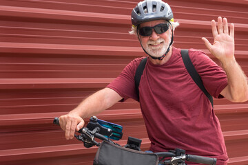 Senior cyclist in outdoor excursion with electric bicycle resting against a red metal panel looking...
