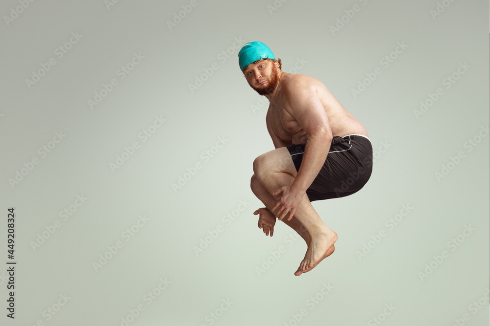 Wall mural Funny cheerful swimmer. Cute red-headed man in red swimming shorts posing isolated on gray studio background. Concept of sport, humor and body positive.