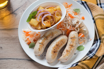 White plate with pan fried munich sausages, sauerkraut and potato salad, elevated view on a brown wooden background