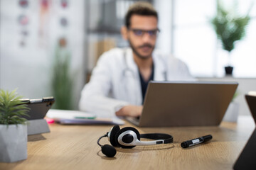 Obraz premium Close up view of headset with microphone and pen on the wooden desk. Blurred view of male Arabian doctor in glasses, focused on his work or online consultation, typing on laptop
