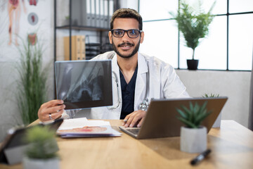 Fototapeta na wymiar Making a diagnosis concept. Portrait of young qualified male Asian Indian doctor, sitting in office, holding skull xray image, working on laptop computer, smiling at camera