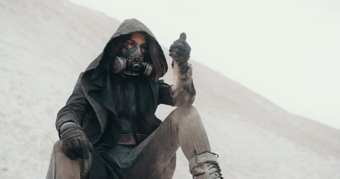 Post apocalyptic female wanderer wearing grunge hooded mantle gas mask hand pouring sand while sitting in desert. Steampunk cyberpunk soldier. Dead world concept, future survivor after nuclear war