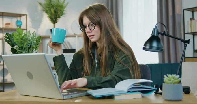 Attractive positive confident smart modern female student in glasses sitting at her workplace at home and working over university home task on computer,drinking tea
