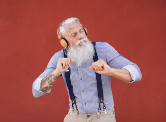 Senior hipster man dancing outdoor while listening music with wireless headphones with red...