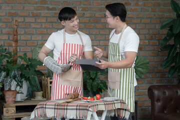 Happy Asian young LGBT gay couple with apron cooking together. Handsome man mixing salad dressing and his boyfriend thumb up to him and using tablet in kitchen at home.