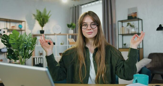 Camera shooting on lovely happy smiling young long-haired woman in casual clothes which meditating with closed eyes at her workplace in home office