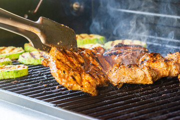 Turning juicy pork steak and grilled zucchini slices prepared on a modern garden grill