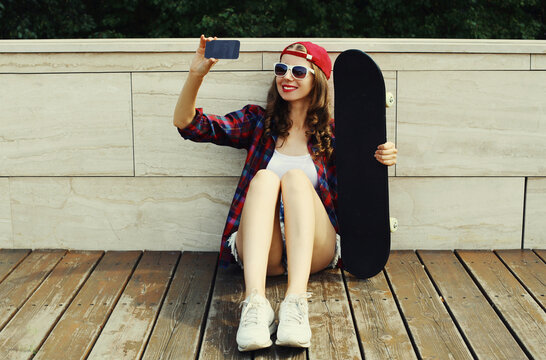 Teenager girl sitting with skateboard taking a selfie by smartphone wearing a baseball cap in the city