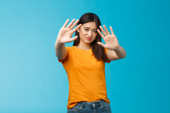 Asian moody displeased silly timid girl asking turn off light not photograph her, raise hands block, cover face step back and cringe disappointed, standing blue background upset reluctant