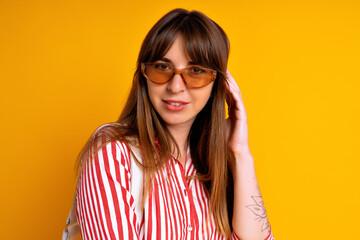 Fototapeta premium Cheerful pretty brunette woman wearing striped red dress, beige sunglasses and white bag, fashionable mood, studio yellow background.