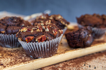 Healthy gluten free chocolate muffin in a baking paper cup with pecan nuts and cocoa powder. Homemade, freshly baked delicious cupcakes in the background on a wooden serving board. Nutrition concept