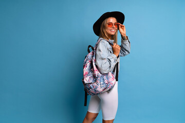 Stylish hipster blonde woman posing at studio blue background. Trendy casual tourist outfit,...