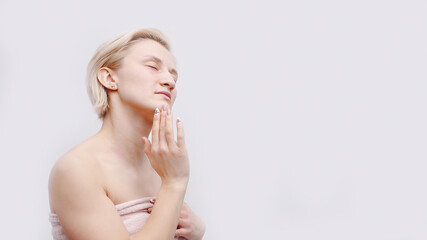 A beautiful blonde model with a towel wrapped around her body. Hand touching her face posing for the camera. Isolated girl over white background studio shot. Spa concept.