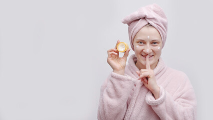Attractive girl holding a small jar of moisturizing skin cream. Applying cream on her face. Finger on lips gesture. The girl wearing a head towel and bathrobe. Concept of moisturizing skin and