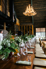 a large wooden table with serving, glasses, green rag napkins and compositions of fresh flowers. White flowers. A room with stone walls. Chandelier made of animal horns. holiday concept.