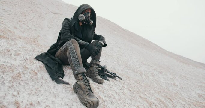 Wide angle post apocalyptic female in grunge hooded mantle and protective gas mask sitting on hill in dead valley desert. Steampunk cyberpunk soldier with gun. Future warrior after nuclear war