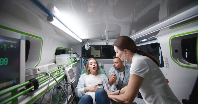 Caucasian young husband supporting pregnant wife in ambulance car during labor and taking selfie photo. Man breathing and holding woman hand when in childbirth. On way to hospital with doctor.