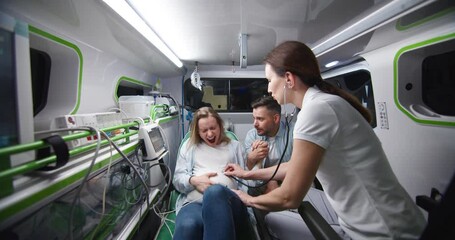 Caucasian husband supporting pregnant wife in ambulance car during labor and taking selfie photo. Man breathing and holding woman hand. Female in childbirth is angry and tired. Medic helping.
