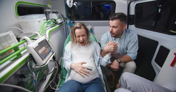 Caucasian young husband supporting pregnant wife in ambulance car during labor and taking selfie photos. Man breathing and holding woman hand when in childbirth. Making photo on way to hospital.