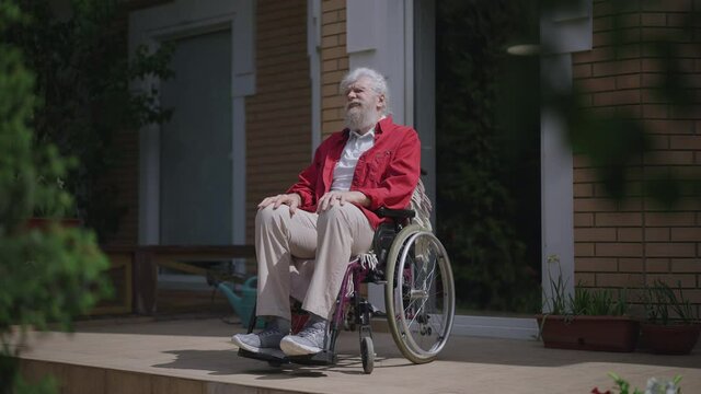Wide shot of old disabled Caucasian man in wheelchair enjoying sunny summer day outdoors on house porch. Portrait of thoughtful elderly male retiree looking around thinking. Slow motion