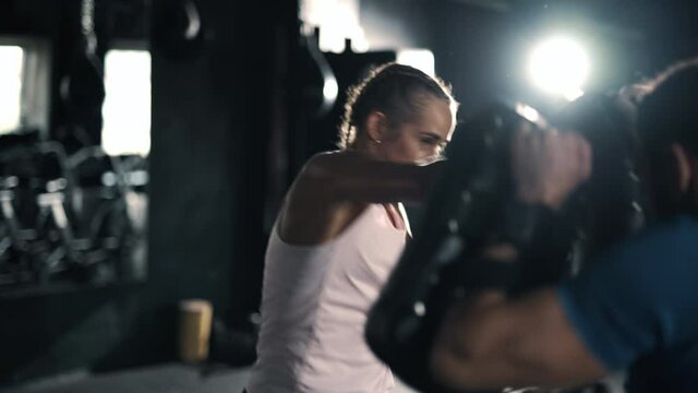 A athlete woman fighter is training punches focus mitts hands and feet in the dark gym. A kickboxer trainer is teaching self defense. Maximum focus and strength. Concept of health and sports.