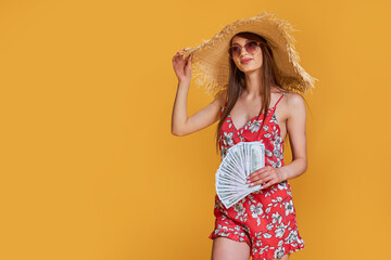 Woman wearing summer straw hat, dress holds a wad of money in her hands and posing on yellow background