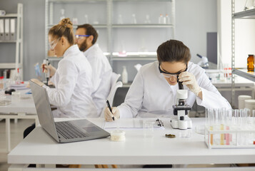 Woman scientist looking through microscope making analysis writing notes during chemical experiment. Research group working in modern well-equipped medical or biochemistry laboratory