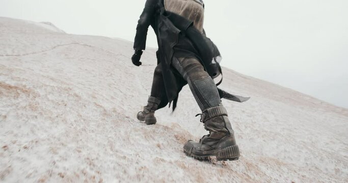 Close-up following shot of female legs stepping. Wide angle post apocalyptic female in futuristic grunge black clothes feet climbing hill in dead desert. Cyberpunk future warrior survivor after war