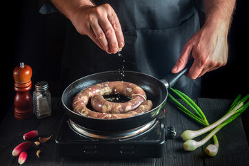 Professional chef adds salt to pan with raw meat sausage. Preparation for cooking sausages in kitchen of restaurant or cafe on table with vegetables and spices.