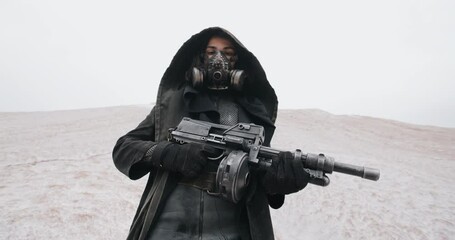 Slow motion post apocalyptic woman in black hooded mantle and grunge protective gas mask posing with gun standing on hill in white dead desert. Cyberpunk future warrior female survivor after war