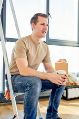 Side View Portrait Of Handsome Caucasian Guy In Casual Wear Having Rest After Home Renovation Sitting On Stepladder, Holding Cup Of Coffee, Relaxing