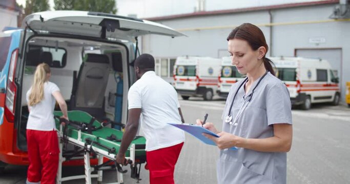 Female doctor filling in medical form and registering. Outdoor. Mixed-races doctors or nurses working at ambulance on background. Portrait of woman medic looking at camera and writing on document.