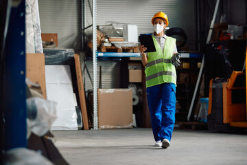 Female worker with face mask uses digital tablet while walking through distribution warehouse.