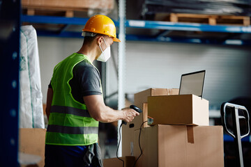 Male worker scans boxes while using laptop and working in warehouse during COVID-19 pandemic.