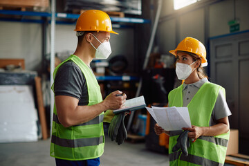 Two workers with protective face masks cooperate while going through delivery schedule in distribution warehouse.