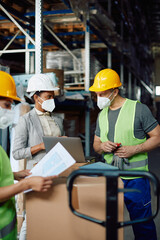 Warehouse worker and black female manager wear face masks while working on laptop at distribution compartment.