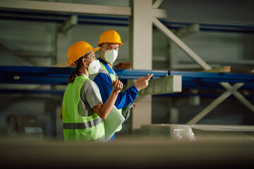 Female worker and her colleague wear face masks while talking and walking through distribution warehouse.