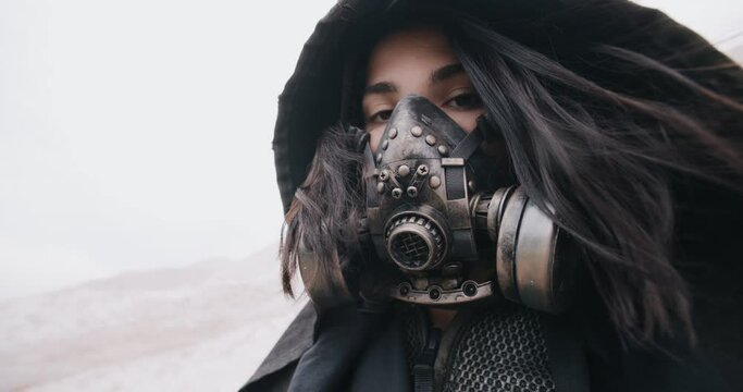 Wide angle portrait post apocalyptic brunette woman wearing grunge hooded mantle protective gas mask in white hilly desert. Stalker concept, cyberpunk future warrior female survivor after nuclear war