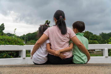 Back view of loving Asian mother hug her kids sitting on bridge, caring black mom embrace child, relaxing looking to black cloud, parent comfort child caressing.