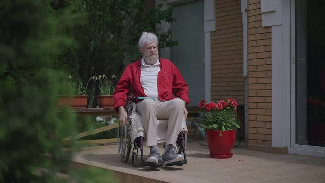 Senior Caucasian disabled man rolling wheelchair on sunny summer porch leaving. Wide shot portrait of confident retiree invalid outdoors in backyard at home. Slow motion