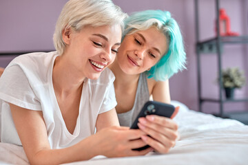 Cheerful lesbian couple having sun on bed using smartphone, watching interesting video.Cute Caucasian short head ladies spending nice time together at home, at weekends. Copy space, portrait