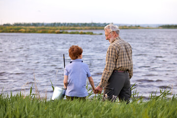 Fototapeta premium Rear View On Positive Grandfather And Grandson In Casual Outfit Going To Fish On Riverside in Countryside, Holding Hands Together. Family, Generation, Summer Holidays And People Concept. Copy Space